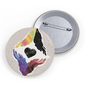 May include: Two round pinback buttons. One button features a design of four colorful hands surrounding a black heart. The other button is silver. The hands are in shades of blue, red, yellow, and black.