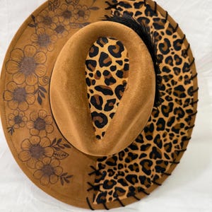 May include: A brown suede fedora hat with a black and brown leopard print trim and a floral design on the other side. The hat has a black feather accent.