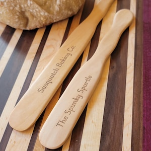 Peut inclure: Deux couteaux à beurre en bois avec du texte gravé. Le couteau du haut porte l'inscription "Sasquatch Baking Co." et le couteau du bas porte l'inscription "The Spunky Spurtle". Les couteaux sont sur une planche à découper en bois rayée.