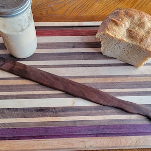 Peut inclure: Une planche à découper en bois avec un motif rayé dans les tons de brun, violet et blanc. Une miche de pain et un bocal en verre avec un couvercle blanc sont sur la planche. Un couteau à pain en bois est également sur la planche.
