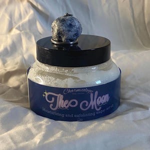 May include: A jar of sugar scrub with a dark blue label featuring the text "Theo Moon" and "Moisturizing and exfoliating sugar scrub." The jar has a black lid and is topped with a blue and silver decorative sphere.