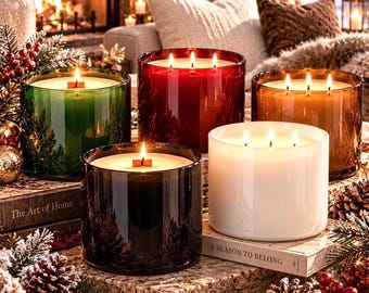May include: Five cylindrical glass candles in green, red, amber, black, and white, each with multiple lit wicks. The candles are arranged on books titled The Art of Home and A Season to Belong, surrounded by pinecones and festive holiday decor.