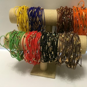 Puede incluir: Exhibición de pulseras de cuentas coloridas en amarillo, azul, marrón y naranja. Las pulseras están adornadas con pequeñas cuentas doradas y están dispuestas en un expositor beige. Otras pulseras son verdes, rojas, negras y marrón oscuro.