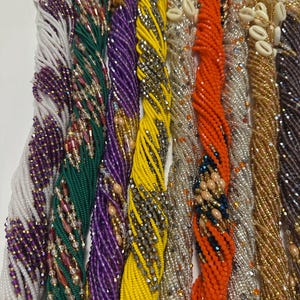 May include: A collection of beaded necklaces in various colors, including white, green, purple, yellow, orange, silver, and brown. Some necklaces feature shell accents. The beads are tightly strung, creating a textured appearance.