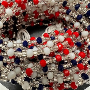 May include: A close-up of a beaded necklace with red, white, and blue beads. The beads are arranged in a pattern, and the necklace is made of multiple strands.