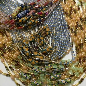 Puede incluir: Una colección de collares de cuentas en varios colores y diseños. Los collares presentan una mezcla de cuentas de vidrio, semillas y decorativas en tonos azules, rosas, dorados, verdes y naranjas. Algunas cuentas tienen patrones florales y geométricos.