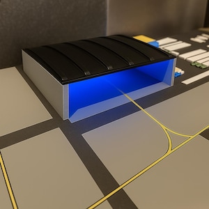 May include: A model airport terminal with a black corrugated roof and a bright blue interior. The terminal has a silver frame and sits on a gray surface with yellow and white markings, resembling an airport runway.