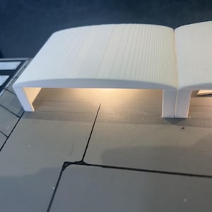 May include: A white, curved architectural model with a light source underneath. The model has a rectangular base and a curved top with vertical lines. The model is on a gray surface.