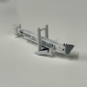 May include: A small, light gray 3D-printed model of a bridge. The model has a detailed design with vertical supports, a textured deck, and a dark gray section at one end. The bridge is set against a plain white background.