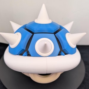 May include: A blue and white spiky shell prop, reminiscent of a video game character. The shell features a segmented blue top with white spikes and a white base. The prop is designed to be worn or displayed.