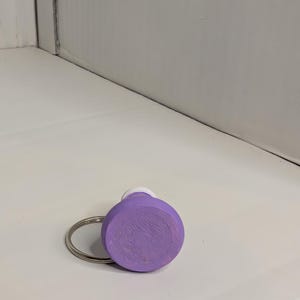 May include: A round, lavender-colored wooden keychain with a silver metal ring. The keychain has a white top and a textured surface. The background is a white surface and a white wall.