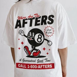 A girl with dark hair wearing a white shirt. She is facing away from the camera. On the back of the shirt is a cartoon Eight Ball and the text “where are the AFTERS” “A guaranteed good time” “CALL 1-800-AFTERS”