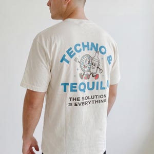 Puede incluir: Camiseta color crema con un gr&aacute;fico en la espalda que presenta una bola de discoteca y una botella de tequila de dibujos animados. El texto dice "TECHNO & TEQUILA" y "THE SOLUTION TO EVERYTHING" en azul. La camiseta tiene mangas cortas.