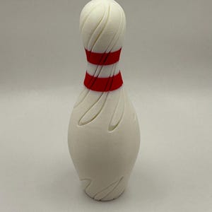 May include: White bowling pin with red stripes around the middle. The pin has a textured surface.