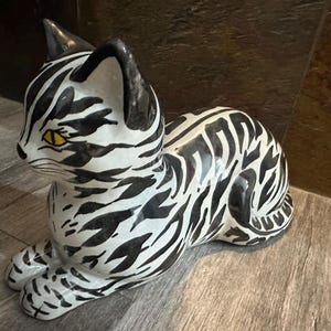May include: A black and white ceramic cat figurine with a yellow eye. The cat is lying down with its head turned to the side. The figurine is painted with a zebra-like pattern.