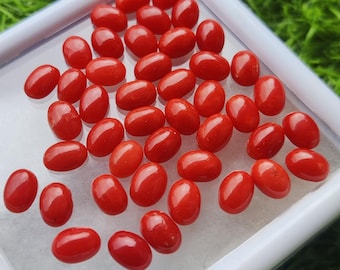 May include: A collection of oval-shaped, polished red gemstones. The gemstones are displayed in a white tray against a backdrop of green grass. The stones have a glossy finish and vary slightly in size.
