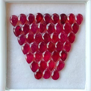 Peut inclure: Une collection de 35 pierres précieuses en forme de poire de couleur rouge rubis disposées en forme de triangle. Les pierres précieuses sont présentées dans une boîte blanche.