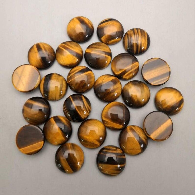 May include: A collection of twenty-one round tiger's eye gemstones. The stones are polished, displaying a mix of golden brown and dark brown bands. The gemstones are arranged on a light gray surface.