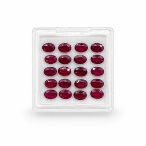 Puede incluir: Una caja de plástico transparente llena de dieciocho rubíes rojos oscuros de corte ovalado. Las piedras preciosas están dispuestas en un patrón de cuadrícula sobre un fondo blanco. Los rubíes tienen una superficie pulida y reflectante, y la caja es transparente.