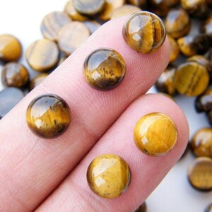 May include: Close-up of several polished tiger's eye gemstone beads. The beads are a mix of golden brown and dark brown, with a chatoyant effect. Some beads are on a finger, while others are scattered in the background. These beads are suitable for jewelry making.