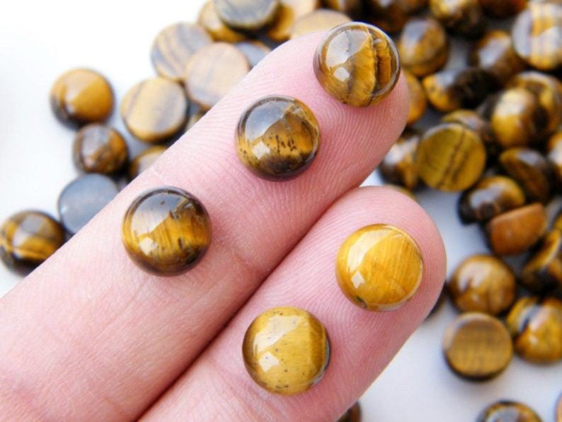 May include: Close-up of several polished tiger's eye gemstone beads. The beads are a mix of golden brown and dark brown, with a chatoyant effect. Some beads are on a finger, while others are scattered in the background. These beads are suitable for jewelry making.