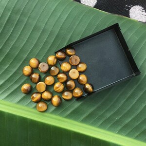 May include: A collection of polished tiger's eye gemstones, each with a chatoyant effect, displayed on a green leaf and a black tray. The gemstones have a warm, golden-brown color with bands of light.