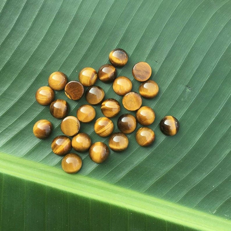 May include: A collection of polished tiger's eye gemstones, each with a rounded shape and a warm, golden-brown color with streaks. The stones are scattered on a large, green leaf, creating a natural and earthy aesthetic. These gemstones are often used in jewelry.