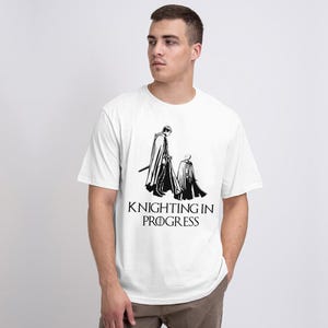 Op de afbeelding: Wit T-shirt met een zwarte afbeelding van twee figuren in mantels, een met een zwaard, en de tekst "Knighting in Progress" eronder. Het T-shirt heeft een ronde hals.