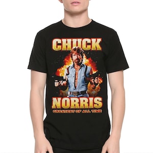 Könnte beinhalten: Schwarzes T-Shirt mit einer Grafik von Chuck Norris, der zwei Waffen vor einem feurigen Hintergrund hält. Der Text "CHUCK NORRIS" steht über dem Bild, darunter "GREATEST OF ALL TIME".
