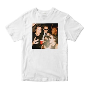 May include: White t-shirt featuring a vintage-style photograph of four people laughing and holding drinks. The image has a warm, retro aesthetic. The t-shirt is a classic crew neck style.