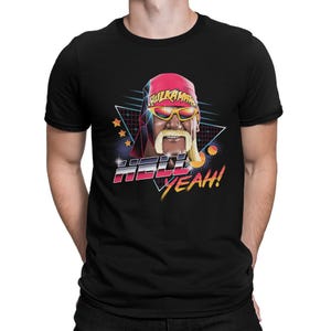 Può includere: T-shirt nera con grafica retrò di un wrestler con bandana rossa e occhiali da sole. Il design include il testo "Hulkamania" e "HELL YEAH!" in un font colorato e vivace.