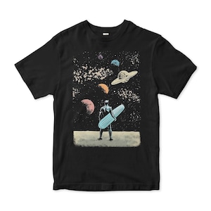 May include: Black t-shirt with a space-themed graphic. The design shows a figure holding a surfboard, with planets and a UFO in the starry background. The shirt is made of a soft material.