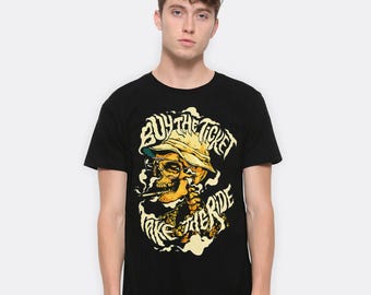 May include: Black t-shirt featuring a skull wearing a hat and smoking a cigarette. The graphic includes the text "BUY THE TICKET TAKE THE RIDE" in a stylized font, with yellow and white accents.