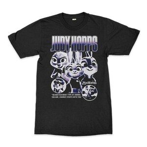 Pu&ograve; includere: T-shirt nera con un disegno di Judy Hopps di Zootropolis. Il design include il testo "JUDY HOPPS" in viola e bianco, e la citazione "NO MATTER WHAT TYPE OF ANIMAL YOU ARE, CHANGE STARTS WITH YOU." La parola "ZOOTOPIA" &egrave; anche visibile.