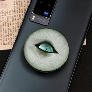 May include: A black smartphone with a rectangular camera module featuring a Zeiss logo, adorned with a circular, textured olive-green phone grip. The grip displays a realistic, narrow-pupil eye with a vibrant green iris centered on its surface.