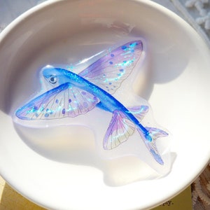 以下が含まれることがあります： 青と紫の空飛ぶ魚の形をした樹脂アート作品。イリデセントグリッターのアクセントがあり、白いボウルに入っています。魚には詳細な翼とひれの模様があります。アートワークはソフトでぼやけた背景に設定されています。