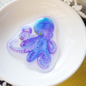 May include: A blue and purple octopus-shaped resin art piece with glitter accents, set in a clear, irregular shape. The octopus has a yellow eye and is displayed in a white ceramic bowl. The artwork is a unique, ocean-themed decorative item.