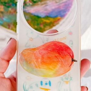 May include: Clear phone case featuring a vibrant, three-dimensional mango design. The case has a circular cutout for the camera and is decorated with whimsical sketches of a dog, cat, and other playful elements.