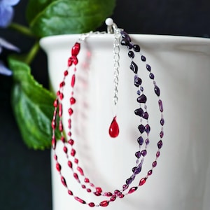 Op de afbeelding: Een gelaagde ketting met rode en paarse kralen. De ketting heeft een zilveren ketting en een enkele rode druppelhanger. De kralen zijn van verschillende vormen en maten, wat een uniek ontwerp creëert. De ketting wordt gepresenteerd tegen een witte achtergrond.