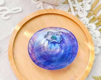 Può includere: Una rappresentazione artistica dettagliata di un mirtillo, con sfumature di blu e viola, poggia su un piccolo piatto rotondo di legno. Il mirtillo ha un aspetto strutturato e un piccolo accento verde. Il piatto è su una tovaglia di pizzo bianco.