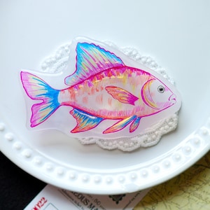 Puede incluir: Un objeto decorativo con forma de pez, con un cuerpo transparente y vibrantes acentos rosas, azules y amarillos. El pez está colocado sobre un plato blanco con un tapete de encaje. Se ve un boleto con el texto "OUS MASTER".