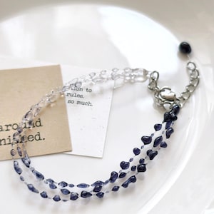 May include: A delicate bracelet featuring three strands of small, dark blue beads that transition to clear beads. The bracelet has a silver-toned chain and clasp. The bracelet is displayed on a white plate, with a small card that reads "around finished."