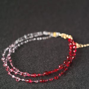 May include: A multi-strand necklace with clear and red glass beads. The beads are arranged in a gradient pattern, starting with clear beads and transitioning to red beads.