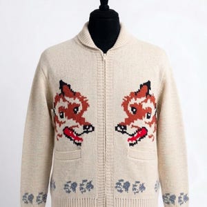 Puede incluir: Una chaqueta de punto beige con cremallera que presenta dos motivos simétricos de cabezas de zorro en el pecho, con detalles de pelaje naranja, blanco y negro y una lengua roja. El dobladillo y los puños tienen un patrón floral gris.