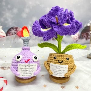 Op de afbeelding: Twee handgemaakte gehaakte items: een paarse bom met een vlam en een paarse bloem in een pot. De bom heeft een bordje met de tekst "Emotional Support Bomb". De bloem heeft een bordje met de tekst "Emotional Support Plant".