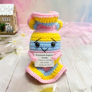 Puede incluir: Un juguete de caramelo de crochet con un cuerpo a rayas rosa, amarillo y azul. El juguete tiene una cara sonriente con ojos negros y una pequeña boca blanca. El juguete sostiene una tarjeta blanca que dice "Emotional Support Candy There is nothing that a little candy can't fix!"