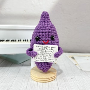 May include: A handmade, purple crochet sweet potato figure with black button eyes and a red stitched smile. It holds a small sign that reads "Emotional Support Sweet Potato". The figure is mounted on a small wooden base.