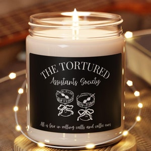 May include: A white candle in a glass jar with a black label that reads "The Tortured Assistants Society" and "All is fair in rolling calls and coffee runs".