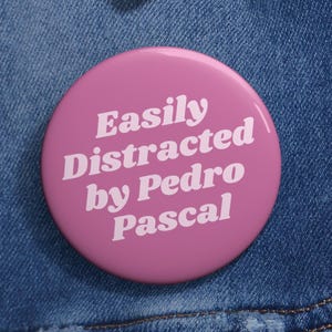 May include: A round, pink button with the text "Easily Distracted by Pedro Pascal" in white lettering. The button is pinned to a blue denim jacket, with visible stitching and metal button details.