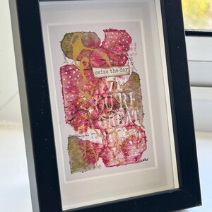 May include: Framed mixed media artwork in a black frame. The artwork features a collage of pink, gold, and beige textures with the words "seize the day" and other text. The piece is signed "LKW".